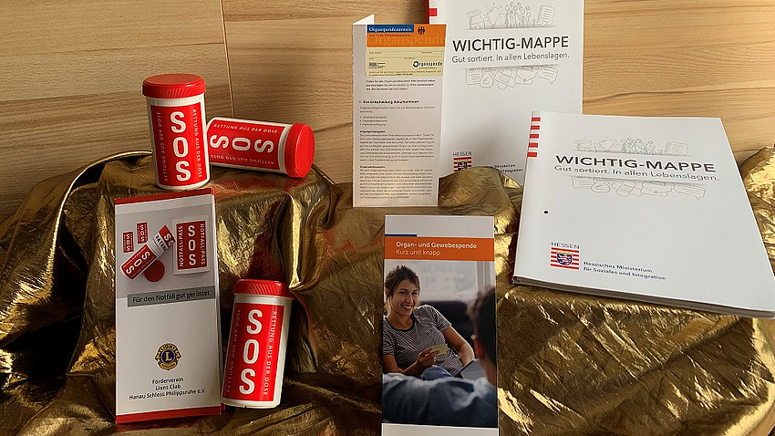 Auf einem Tisch drapiert liegen mehrere rote SOS-Dosen, eine Wichtig-Mappe und ein Flyer mit einem Organspendeausweis.