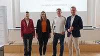 Zwei Frauen und zwei Männer stehen in einem Saal vor einem Podest. Im Hintergrund ist eine Leinwand mit einem darauf abgebildeten Dokument zu sehen.