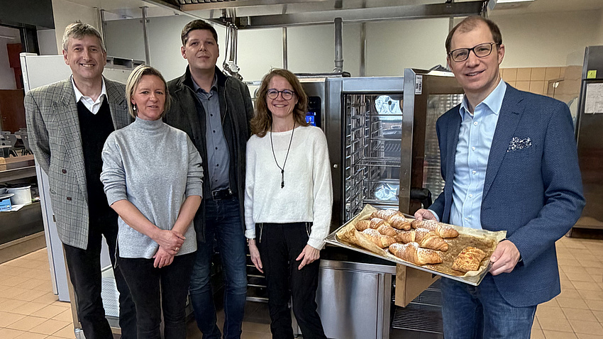 Zwei Frauen und drei Männer stehen in einem Küchenbereich, im Hintergrund sind größere Küchengeräte zu sehen. Einer der Männer hält ein Blech mit Gebäck in Händen.