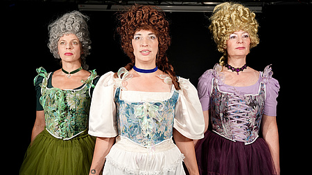 3 historisch kostümierte Frauen halten sich an den Händen.