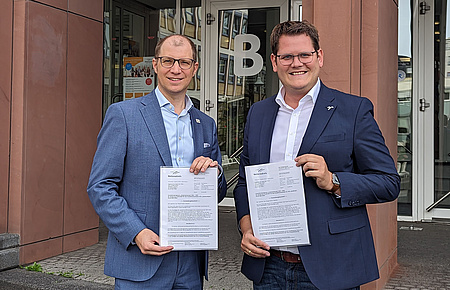 Zwei Männer stehen nebeneinander vor einem Gebäude. Jeder der beiden hält ein Schriftstück in der Hand.