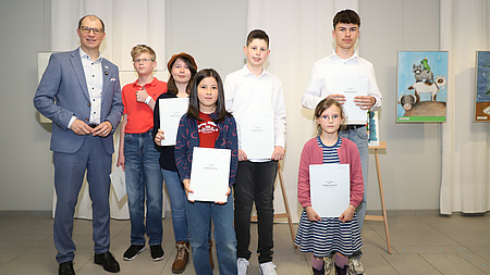 Ein Mann steht gemeinsam mit sechs Kindern und Jugendlichen in einem größerem Raum. Im Hintergrund sind gemalte Bilder an den Wänden zu sehen, und die Kinder und Jugendlichen halten Dokumente in Händen.