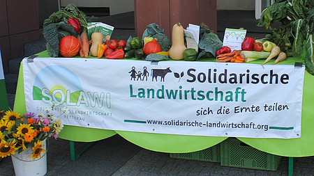 Zu sehen ist ein mit Obst und Gemüse dekorierter Tisch. Daran hängt ein Schild, auf dem "Solidarische Landwirtschaft" steht.
