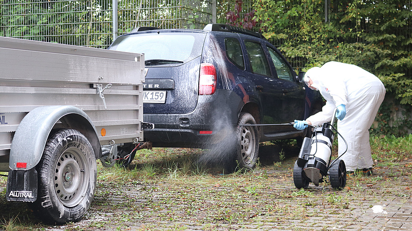 Ein Mann in einem weißen Ganzkörper-Schutzanzug desinfiziert die Reifen eines Autos und eines Anhängers mit einer weißen Flüssigkeit.