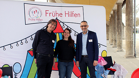 Eine Frau und zwei Männer stehen zusammen in einem Säulengang vor einer großen, bunten Plakatwand mit der Aufschrift "Frühe Hilfen".