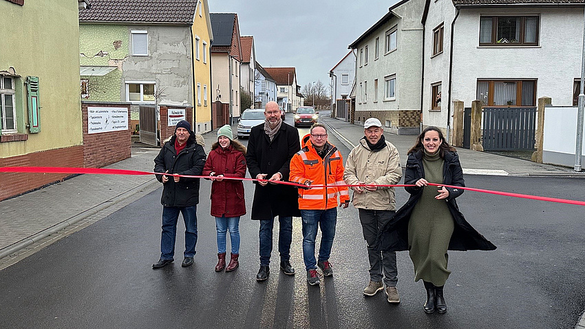 Sechs Männer und Frauen stehen auf einer frisch sanierten Straße. Sie schneiden mit Scheren ein rotes Band durch.