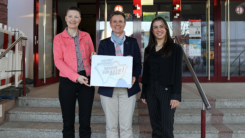 Drei Frauen stehen vor einer Treppe, die zu einem Eingang mit Glastüren führt. Zwei von ihnen halten ein Plakat in die Höhe.