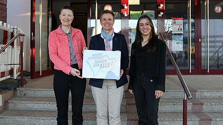 Drei Frauen stehen vor einer Treppe, die zu einem Eingang mit Glastüren führt. Zwei von ihnen halten ein Plakat in die Höhe.
