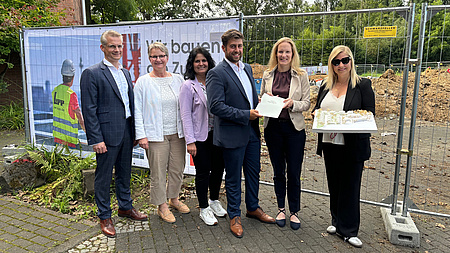 Eine Gruppe von Menschen steht vor einem Bauzaun und hält Dokumente in die Kamera.