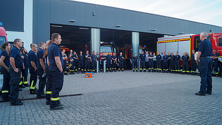 Viele Feuerwehrmänner und Feuerwehrfrauen stehen in einem Halbkreis. Im Hintergrund ist ein Feuerwehrauto zu sehen.