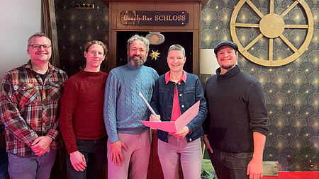 Vier Leute lächeln in die Kamera. Sie zeigen ein Dokument.