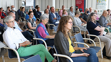 In einem großen Saal sitzen viele Personen in Stuhlreihen hintereinander.
