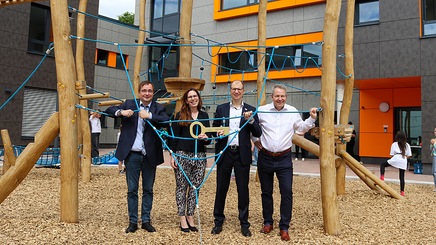 Eine Frau und drei Männer stehen an einem Klettergerüst vor einem großen Gebäude. Einer der Männer übergibt einen großen, goldenen Schlüssel an die Frau.
