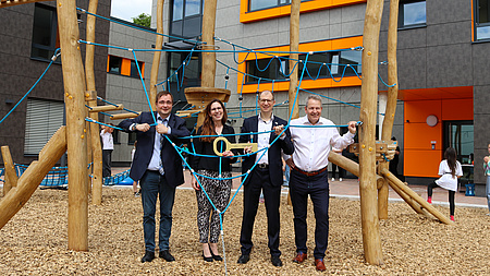 Eine Frau und drei Männer stehen an einem Klettergerüst vor einem großen Gebäude. Einer der Männer übergibt einen großen, goldenen Schlüssel an die Frau.