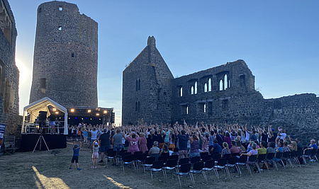Rund 300 Personen sitzen in Stuhlreihen vor einer Bühne. In Hintergrund die Ruinen der Burg Münzenberg