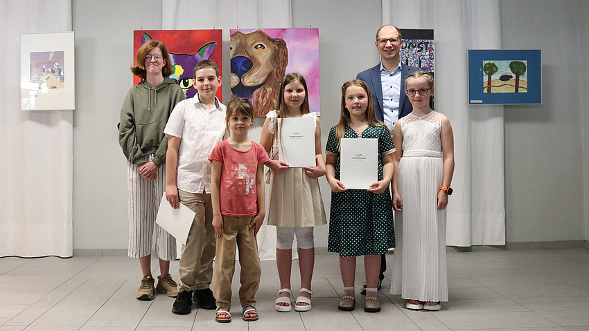 Sieben Menschen stehen vor einer Bildergalerie und halten Urkunden in den Händen.