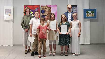 Sieben Menschen stehen vor einer Bildergalerie und halten Urkunden in den Händen.