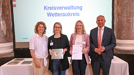 Drei Frauen und ein Mann stehen in einem Saal vor einem Tisch und einer Leinwand. Eine der Frauen hält eine Urkunde in Händen, eine weitere eine Plakette.
