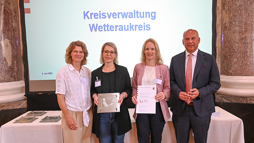 Drei Frauen und ein Mann stehen in einem Saal vor einem Tisch und einer Leinwand. Eine der Frauen hält eine Urkunde in Händen, eine weitere eine Plakette.