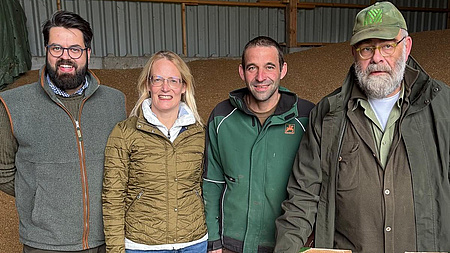 Ein Frau und drei Männer stehen in einer landwirtschaftlich genutzten Halle. Im Hintergrund ist eine große Menge gelagertes Getreide zu sehen.