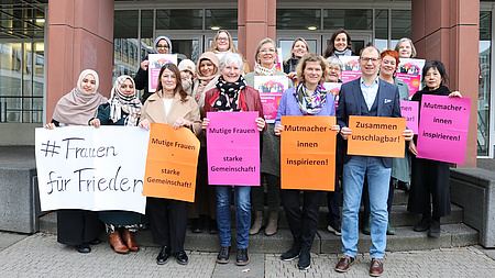 Ein Mann und mehrere Frauen stehen vor und auf der Treppe zu einem Gebäude. Die Personen in der vorderen Reihe haben Plakate mit positiven Ausagen zu Frauen in den Händen