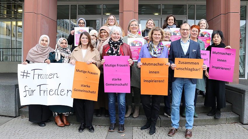 Ein Mann und mehrere Frauen stehen vor und auf der Treppe zu einem Gebäude. Die Personen in der vorderen Reihe haben Plakate mit positiven Ausagen zu Frauen in den Händen