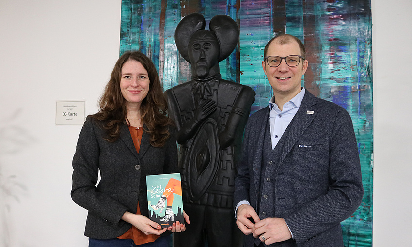 Ein Mann und eine Frau stehen vor einer großen schwarzen Statue. Die Frau hält ein Buch in der Hand. Im Hintergrund an der Wand ein großformatiges abstraktes Gemäde.