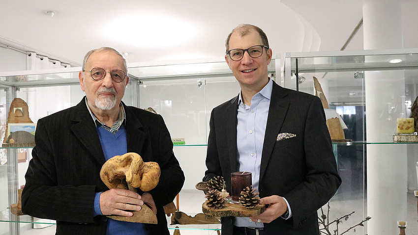 Zwei Männer stehen vor Vitrinen und halten zwei hölzerne Kunstwerke in der Hand.