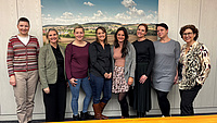 Acht Frauen stehen in einem Büroraum vor einer Wand mit einem Landschaftsfoto.