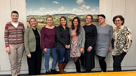 Acht Frauen stehen in einem Büroraum vor einer Wand mit einem Landschaftsfoto.