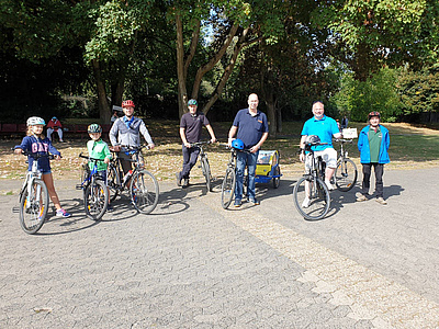 Eine kleine Personengruppe mit Fahrrädern, im Hintergrund eine Wiese mit Bäumen