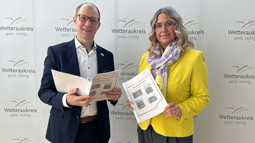 Der Landrat und eine Mitarbeiterin halten den Schulbaubericht in den Händen und blicken in die Kamera.