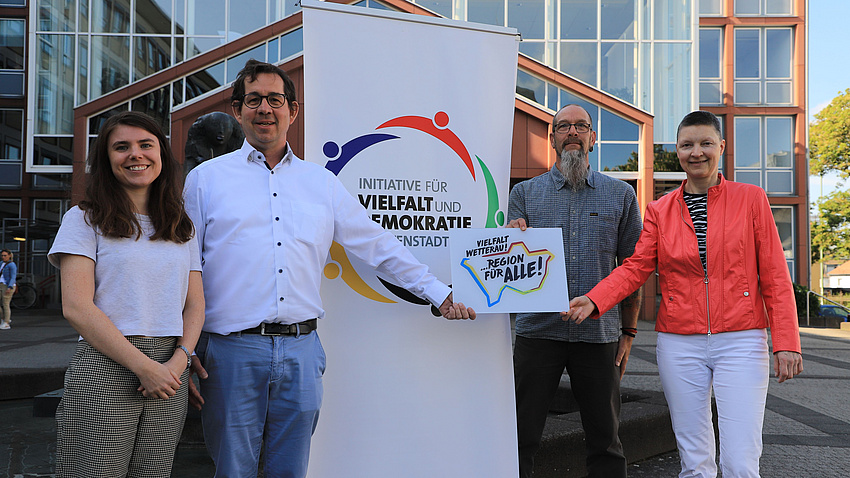 Zwei Frauen und zwei Männer stehen auf einem Platz vor einem Gebäude. Zwischen ihnen steht ein Plakat. Eine der Frauen übergibt einem der Männer ein Dokument.