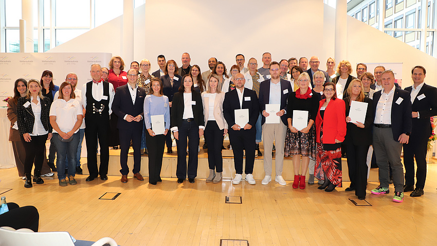 Zahlreiche Frauen und Männer stehen in einem Saal zusammen. Viele von ihnen halten Urkunden in Händen.