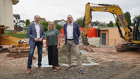 Drei Personen stehen auf einer Baustelle, im Hintergrund sind Container zu sehen und ein Bagger.