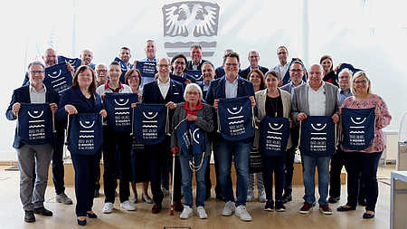 Viele Menschen halten jeweils einen mit einem Logo bedruckten Stoffbeutel in der Hand.