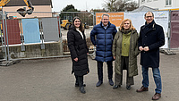 Zwei Frauen und zwei Männer stehen gemeinsam im Freien auf einem Platz. Im Hintergrund sind Gebäude, Baustellenabsperrungen und ein Bagger zu sehen.