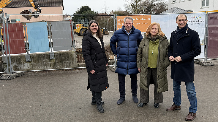 Zwei Frauen und zwei M&auml;nner stehen gemeinsam im Freien auf einem Platz. Im Hintergrund sind Geb&auml;ude, Baustellenabsperrungen und ein Bagger zu sehen.