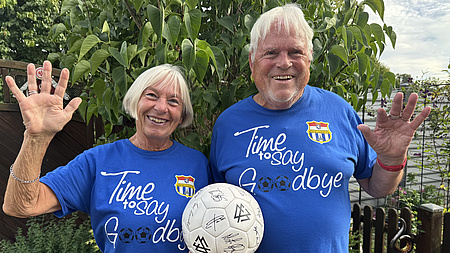 Ein älteres Ehepaar lächelt in die Kamera. Beide halten gemeinsam einen Fußball.