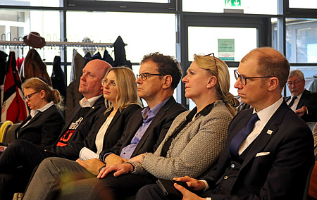 Drei Frauen und drei Männer sitzen nebeneinander in einem Saal. Sie hören einer Person zu, die auf der Bühne spricht.