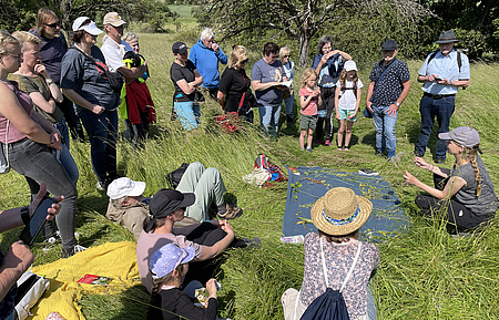 Mehrere Personen sitzen und stehen im Kreis auf einer Wiese. Auf einem großen Tuch in der Mitte liegen Pflanzen. Davor eine Frau, die die Pflanzen erklärt.