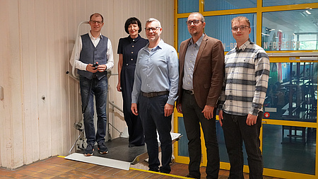 Fünf Personen rund um einen Lift, der an einer Treppe angebracht ist.