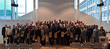Eine große Personengruppe steht in einem großen Saal. Viele der Abgebildeten halten Wimpel in Händen.