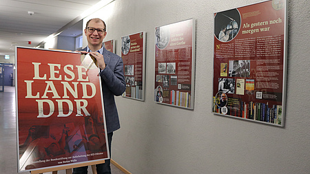 Landrat Jan Weckler steht neben einer Staffelei mit dem ersten Plakat der aktuellen Ausstellung. Im Hintergrund hängen an der Wand die Bilderrahmen mit den Ausstellungsplakaten.
