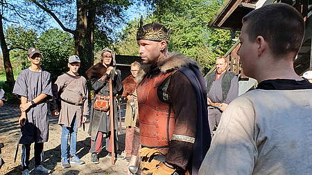 Mehrere Jugendliche in mittelalterlichen Tuniken haben sich um einen als König verkleideten Schauspieler gruppiert.
