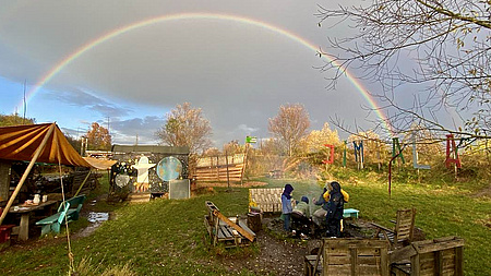 Kinder an einem Feuer im Freien, über ihnen ein Regenbogen