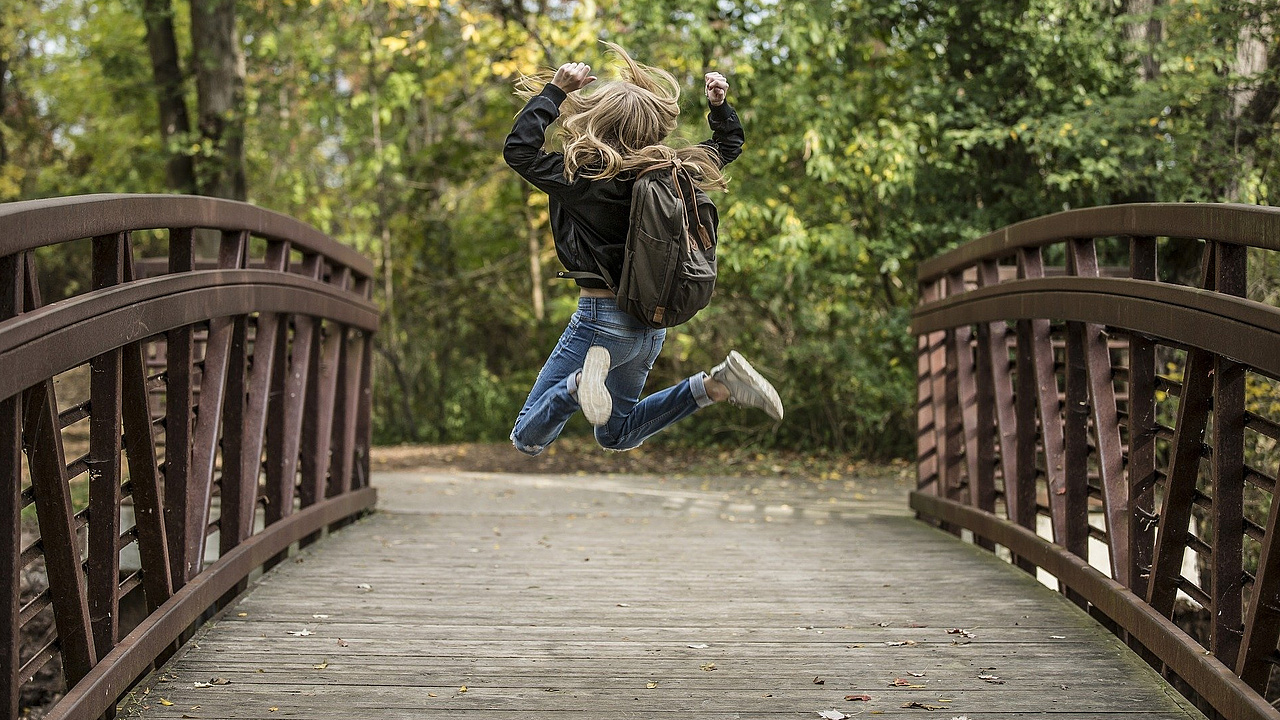 Ein größeres Kind springt freudig über eine Brücke, einen hölzernen Steg, der von der einen Seite des Bachufers zur anderen führt.