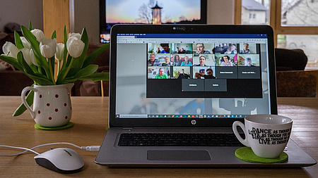 Auf einem Tisch steht ein aufgeklappter Laptop, daneben eine Blumenvase mit Tulpen, eine Computermaus und eine Tasse.