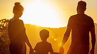 Ein Frau, ein Kind und ein Mann stehen Hand in Hand im Freien und schauen, wie die Sonne hinter einem Berg untergeht.