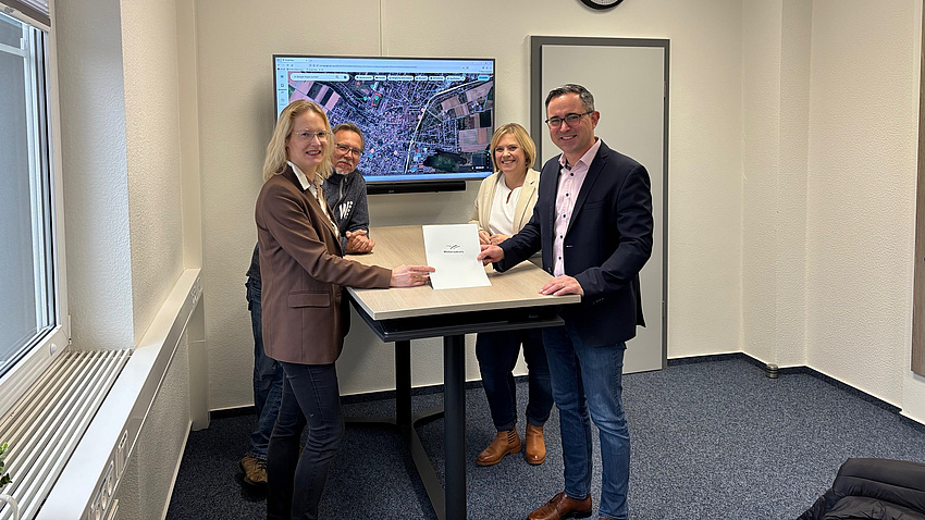 Zwei Frauen und zwei Männer stehen in einem Büroraum an einem Tisch. Eine der Frauen und einer der Männer halten gemeinsam ein Dokument in Händen.